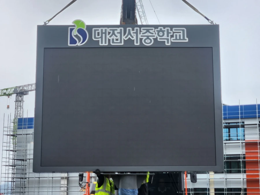 수도권 현장에서 학교 명판이 달린 검은 화면의 전광판을 크레인 줄에 매달아 들어 올린 모습